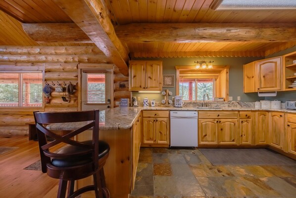 A log cabin kitchen with wooden cabinets, a stone-tiled floor, and a bar area with a chair.