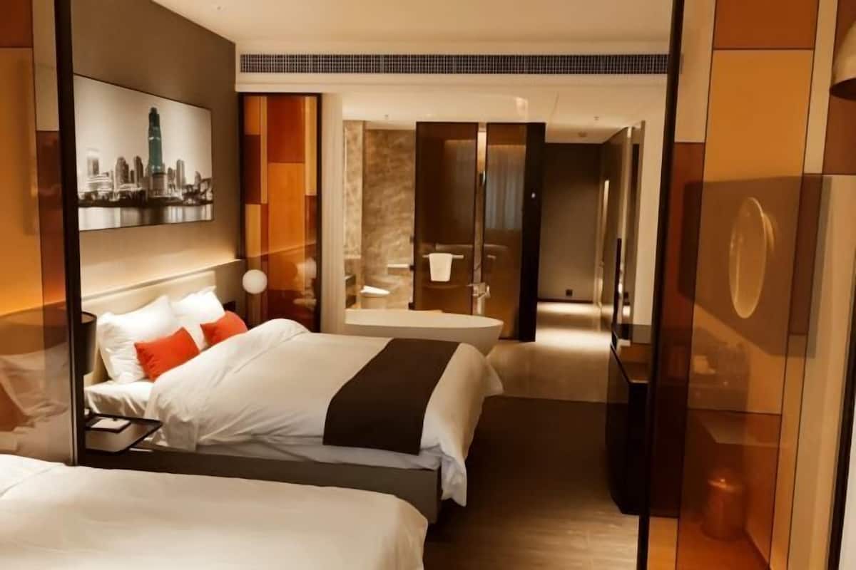 Habitación de hotel con dos camas, un espejo grande, una obra de arte con vista a la ciudad y un baño moderno con separador de vidrio.