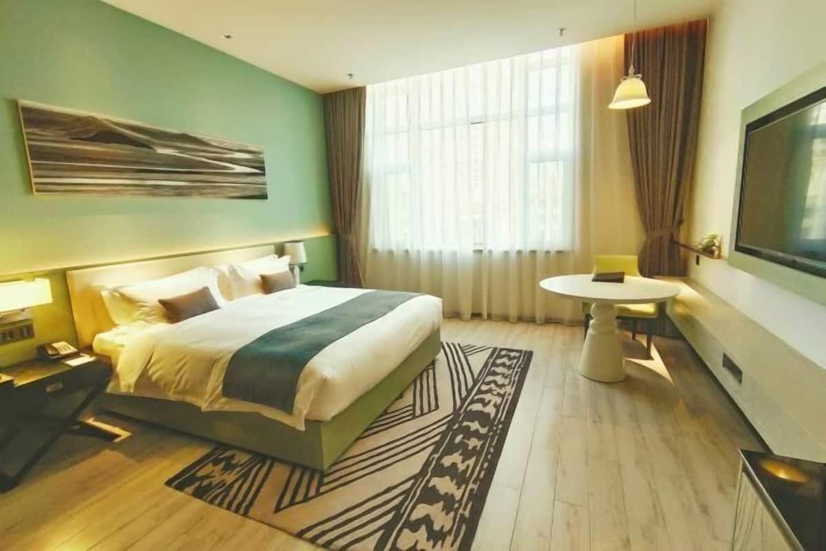 Habitación de hotel con cama, mesitas de noche, una mesa redonda, televisor y un ventanal grande con cortinas.