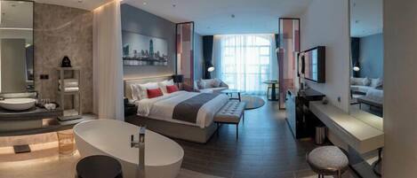 A modern hotel room with a large bed, a separate sitting area, a bathtub, and a television.