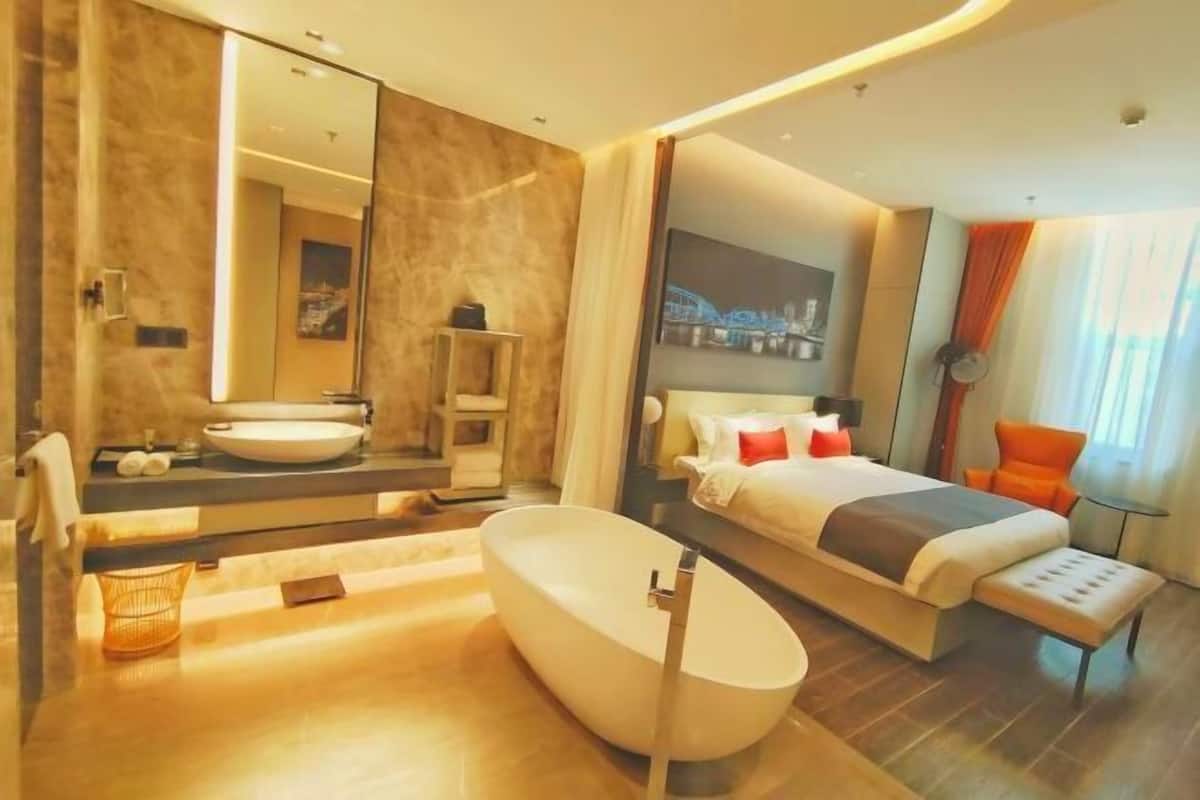Una habitación de hotel moderna con una bañera ovalada grande, una cama doble, una mesita de noche, un espejo y un amplio ventanal con cortinas.