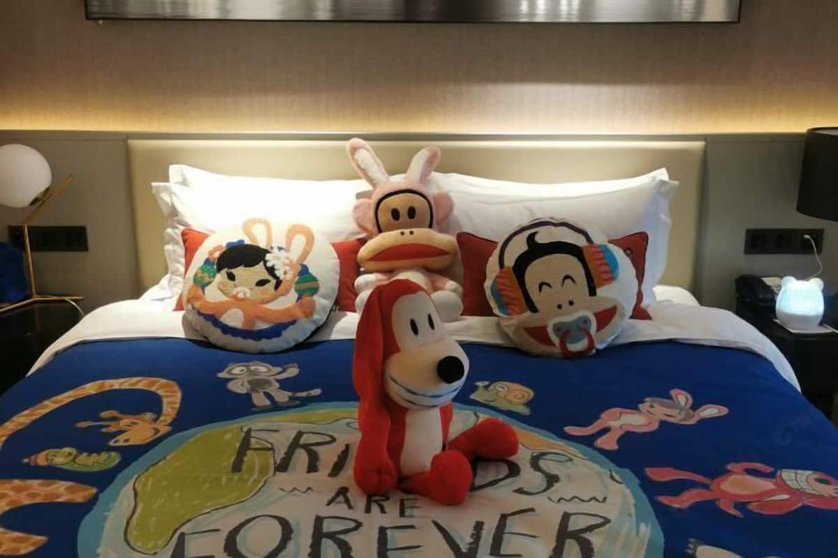 Una cama con almohadas de personajes de dibujos animados, una colcha azul con el mensaje 'FRIENDS ARE FOREVER' y un televisor montado en la pared.