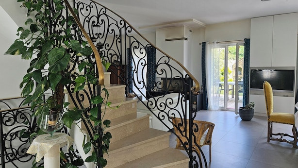 Una escalera con barandillas negras ornamentadas, una planta en maceta y una silla de mimbre.