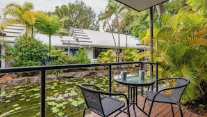 Eine Terrasse mit Tisch und Stühlen mit Blick auf einen Teich mit Seerosen, umgeben von tropischen Pflanzen und einem Gebäude mit weißem Dach.