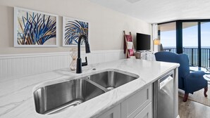 A modern kitchen with a double sink, white marble countertops, and a view of the ocean through large windows.