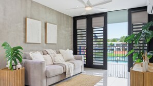 Una sala de estar moderna con un sofá color beige, dos obras de arte enmarcadas, un ventilador de techo y ventanas amplias con vistas a una piscina y zonas verdes.