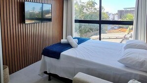A hotel room with a bed, a sofa, a TV mounted on a wooden panel wall, and a large window offering a view of the outside. - Villas do Porto - 212 by La Playa (Ipojuca)
