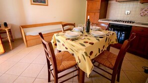 A dining area with a table set for four, a bench, and a kitchen in the background. - Eucalyptus Apartment 3 (Porto Azzurro)