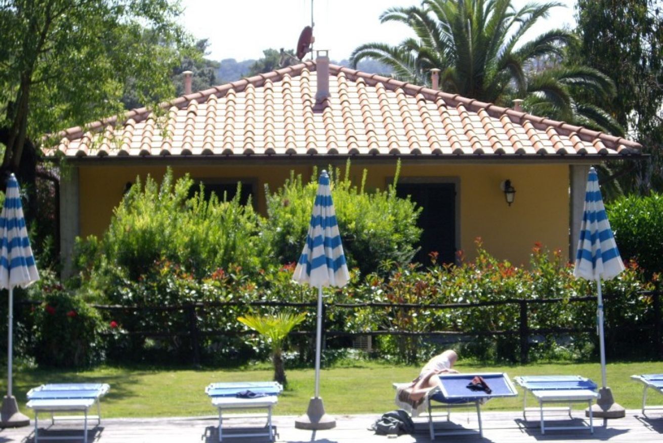 A building with a tiled roof, surrounded by greenery and palm trees. There are lounge chairs and umbrellas in front.