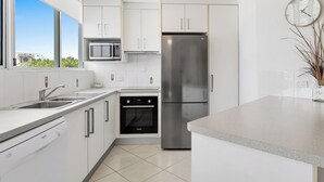 A modern kitchen with white cabinetry, stainless steel appliances, a built-in microwave, and a clock on the wall. - Beachside Apartment (Sunshine Coast)