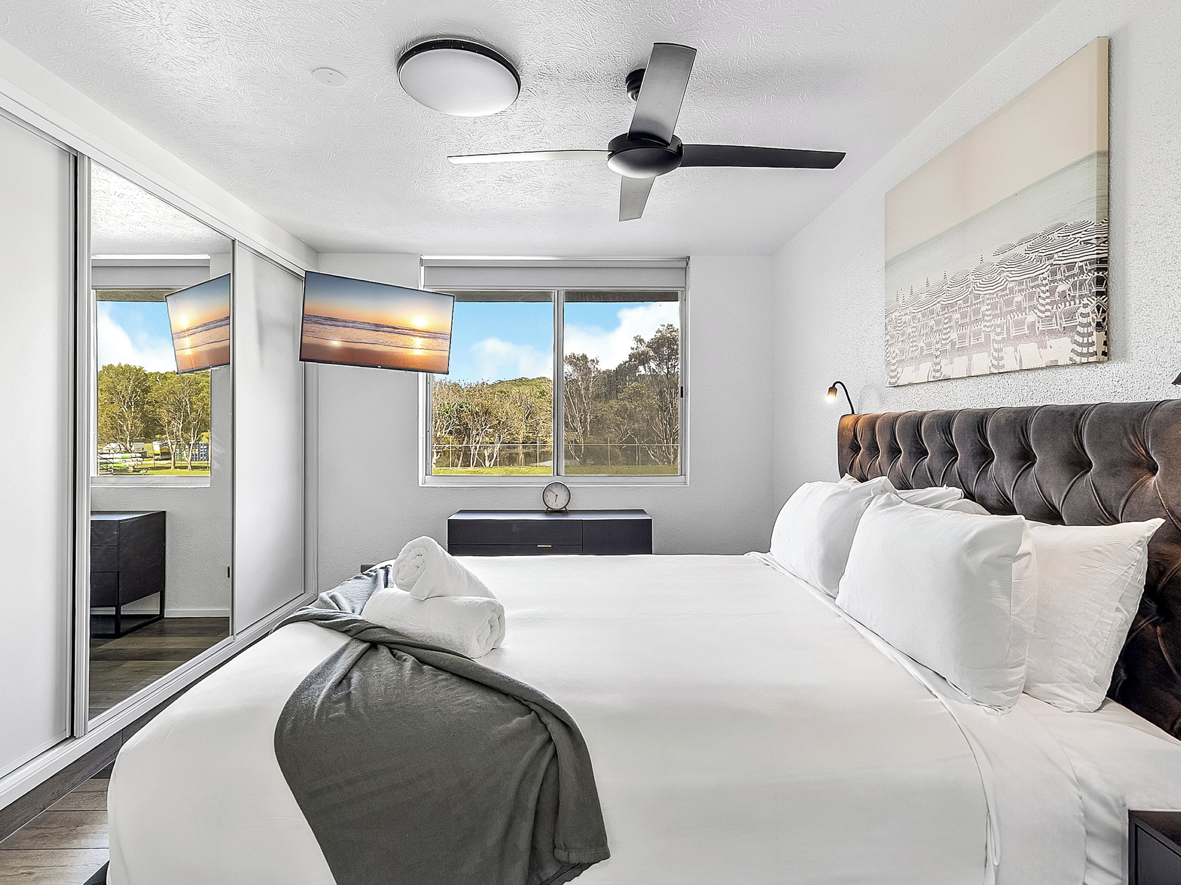 A modern bedroom with a large bed, a ceiling fan, a TV, and a view of the outdoors.