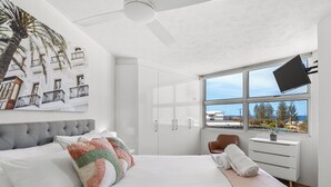 A modern bedroom with a large bed, a ceiling fan, a window with a view, and a flat-screen TV mounted on the wall.
