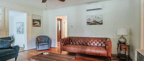 Una sala de estar con un sofá de cuero marrón, un sillón gris oscuro, una mesa de centro de madera y un ventilador de techo.