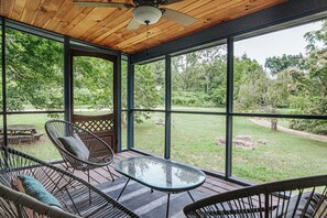 Une terrasse couverte, dotée de chaises en osier et d’une table en verre, et de grandes fenêtres donnant sur une cour verdoyante.