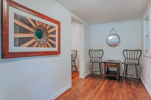 Un couloir doté d’un plancher en bois, d’une œuvre d’art encadrée, de deux chaises, d’une petite table et d’un miroir accroché au mur.