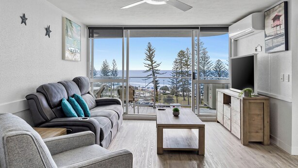 A modern living room with a sofa, a coffee table, and a TV. There is a balcony with a view of trees and a building. - Ocean View Superior Apartment (Alexandra Headland)