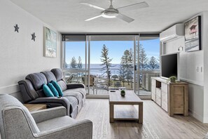 A modern living room with a sofa, a coffee table, and a TV. There is a balcony with a view of trees and a building.