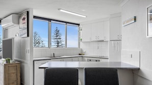 A modern kitchen with white cabinets, a large window, and a dining area with two chairs and a table. - Ocean View Superior Apartment (Alexandra Headland)