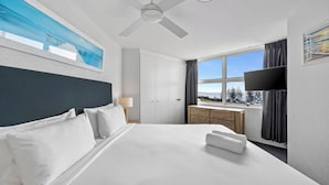 A hotel room with a bed, a ceiling fan, a window with curtains, a TV, and a painting on the wall. - Ocean View Superior Apartment (Alexandra Headland)