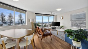 A modern living room with a dining area, large windows, a ceiling fan, a flat-screen TV, and a large potted plant. - Ocean View Superior Apartment (Alexandra Headland)