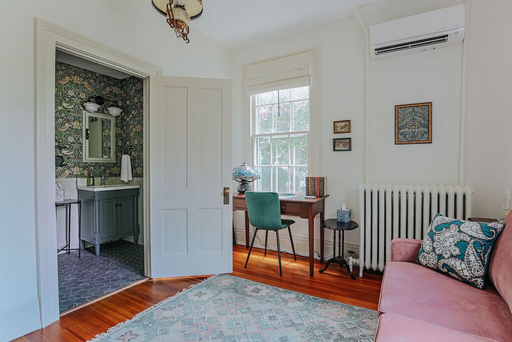 A room with a wooden floor, a pink sofa, a green chair, a small table, a radiator, and a window with a view of trees.