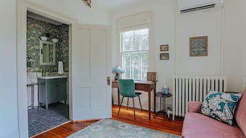 A room with a wooden floor, a pink sofa, a green chair, a small table, a radiator, and a window with a view of trees.