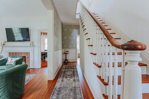Eine Treppe mit weißen Balustern und einem hölzernen Geländer führt zu einer oberen Etage. Im Vordergrund sind ein grüner Sessel und ein Kamin zu sehen.