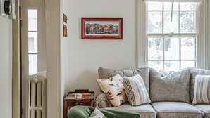 Un salon chaleureux avec un canapé gris, un fauteuil vert et un radiateur. Un tableau encadré est accroché au mur, et une fenêtre laisse entrer la lumière naturelle.