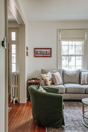 A cozy living room with a grey sofa, a green armchair, and a radiator. A framed picture hangs on the wall, and a window provides natural light.