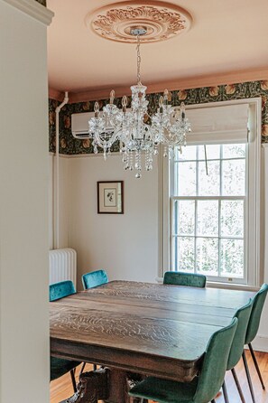 A dining room with a wooden table, green upholstered chairs, a crystal chandelier, a white radiator, and a window with a view of trees. - The Woodward: Walk Downtown, Staunton Mural (Staunton)