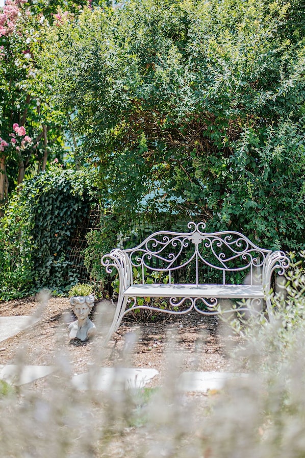A white metal bench in a garden with dense foliage and a statue in the foreground.