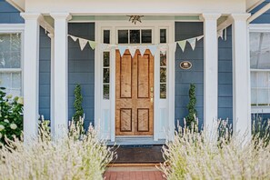 Una casa azul con puerta de madera, columnas blancas y un pequeño jardín con flores blancas.