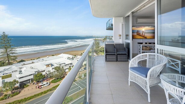 A balcony with a view of the beach and ocean. There are wicker chairs and a table.