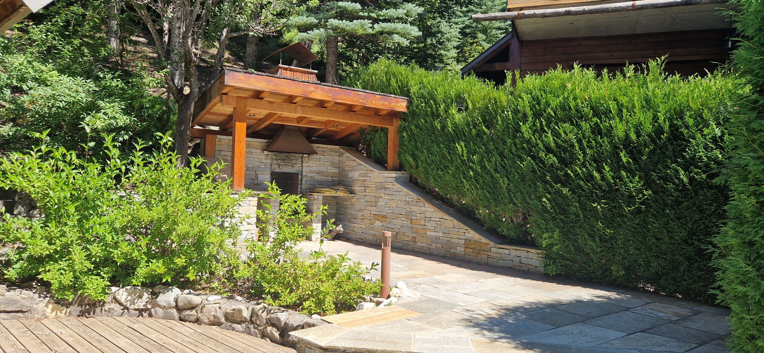Una pérgola de madera con chimenea de piedra, rodeada de abundante vegetación y una escalera de piedra.