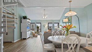 Un vaste espace de vie comprenant une table à manger, des chaises blanches, un ventilateur de plafond et un escalier menant à l’étage supérieur.