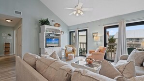 Un salon avec un canapé beige, un meuble de rangement blanc pour les appareils électroniques, un ventilateur de plafond et une vue sur l’extérieur, visible à travers une porte vitrée.