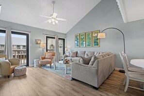 Un salon spacieux avec un grand canapé, une table basse et une table à manger. On y trouve un ventilateur de plafond, une grande fenêtre avec des rideaux et une vue sur un balcon.