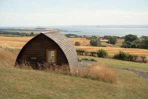 Una cabana de fusta amb teulada corbada, ubicada en un turó herbós amb vista a una massa d'aigua i turons allunyats.