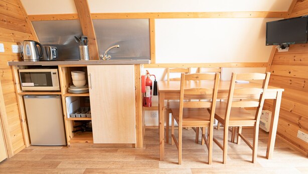 A compact kitchen with a microwave, kettle, and sink. There is a small refrigerator, wooden chairs, and a table. - Rural Cosy Cabin With Beautiful Views Over Fife (Leven)