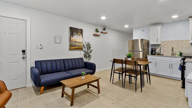 Ett kompakt bostadsutrymme med en soffa, ett matbord, stolar, ett litet köksområde och en dörr.