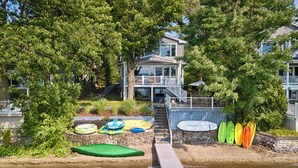 A two-story house with a deck, surrounded by lush greenery. - Lake Escape: The Definition of Lakefront Luxury (Canandaigua)