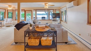 A spacious living room with a large sofa, a coffee table, and a television. There are large windows offering a view of the outdoors. - Lake Escape: The Definition of Lakefront Luxury (Canandaigua)