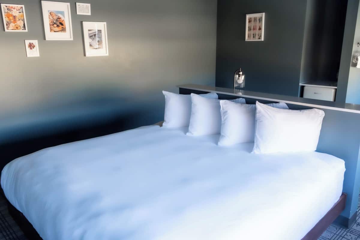 A neatly made bed with white linens in a hotel room with a dark blue wall and a small bedside table.