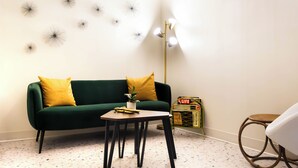 En grön soffa med gula kuddar, ett litet bord med en växt, en golvlampa, en bokhylla och en rund stol.