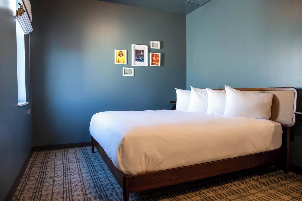 A neatly made bed in a room with a blue wall, a patterned floor, and several framed pictures on the wall.