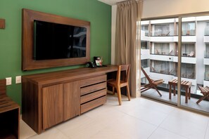 Una habitación moderna con una amplia televisión de pantalla plana, muebles de madera, un escritorio y vistas a un edificio residencial a través de grandes puertas de vidrio.