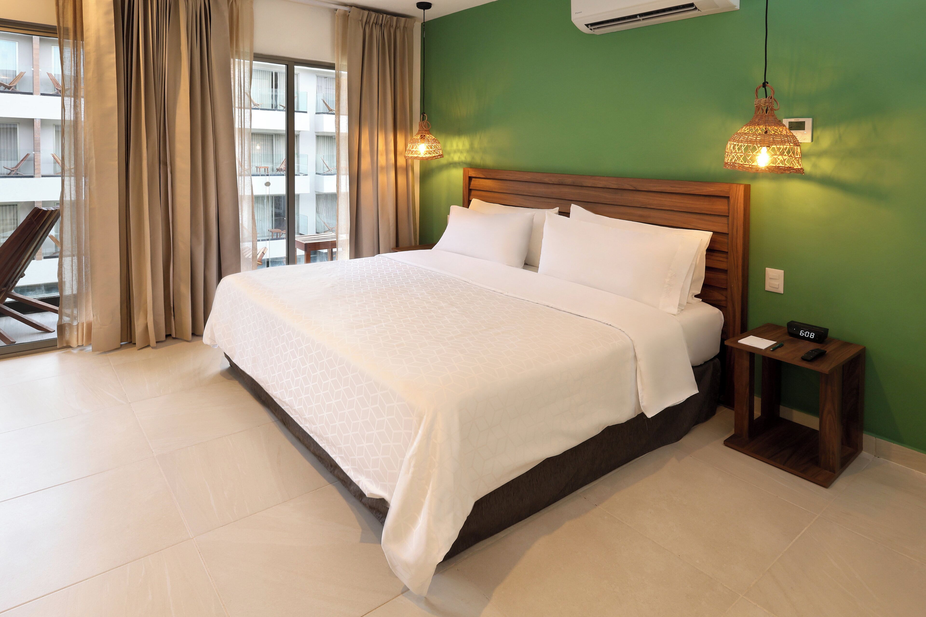 A hotel room with a large bed, a bedside table with a lamp, a window with curtains, and a view of the outside balcony.