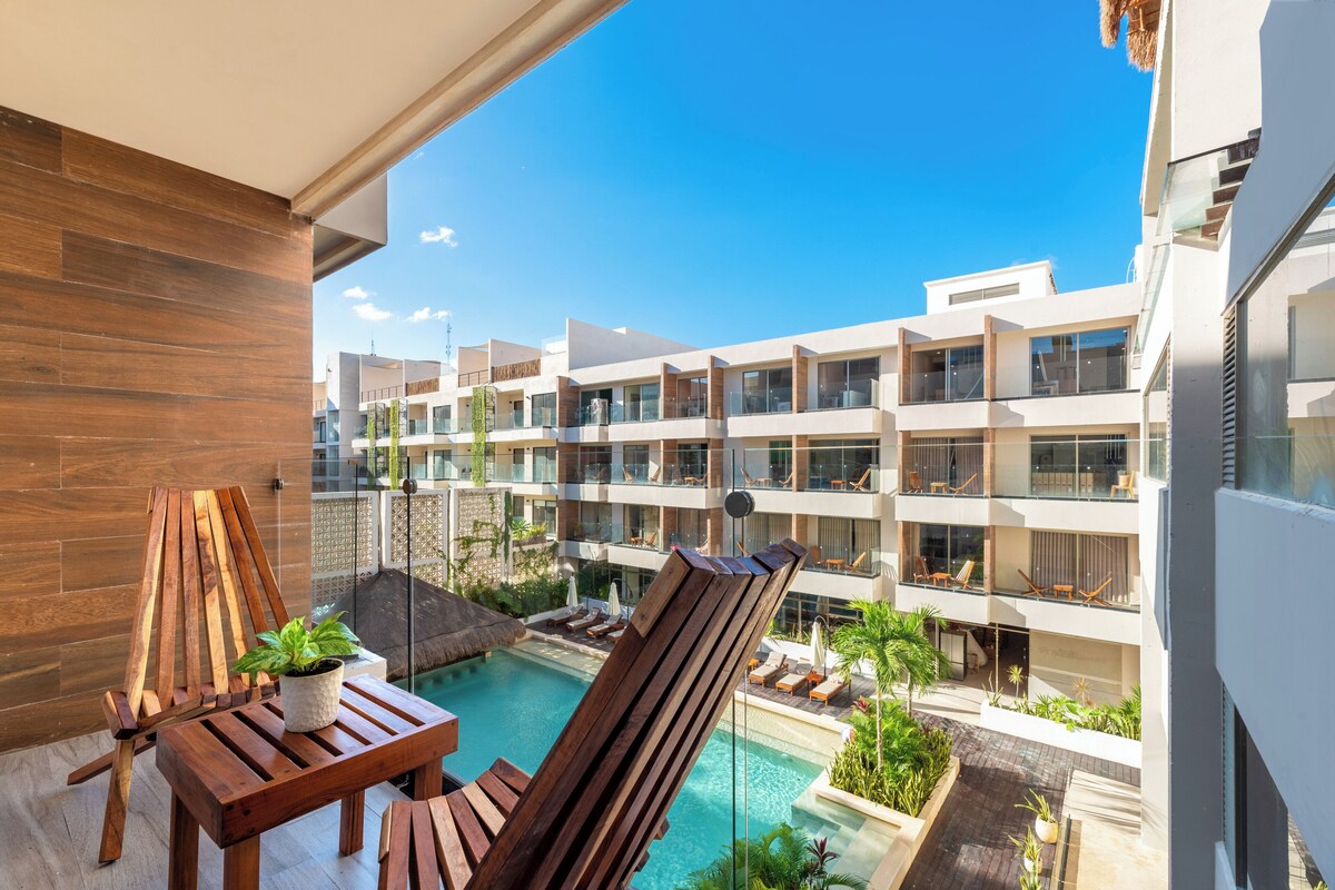 Un balcon moderne d’hôtel, avec une table en bois, une plante en pot et un espace piscine agrémenté de chaises longues et de plantes.