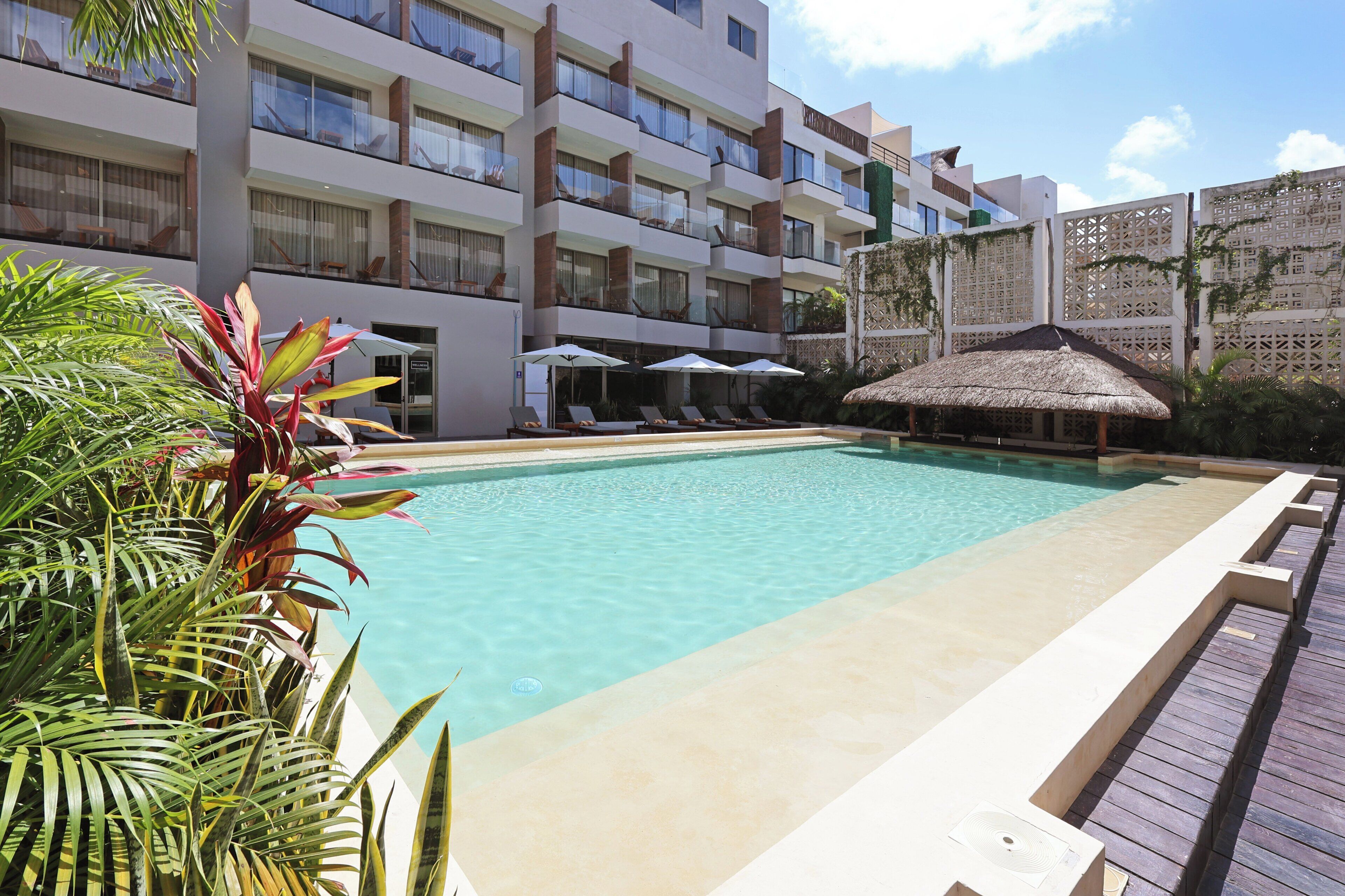 Una piscina con sillones de descanso y sombrillas, rodeada por un edificio de hotel de varios pisos con balcones.