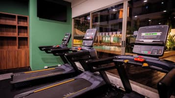 Dans une salle de sport, on trouve trois tapis de course, un mur vert et une grande fenêtre donnant sur un immeuble.